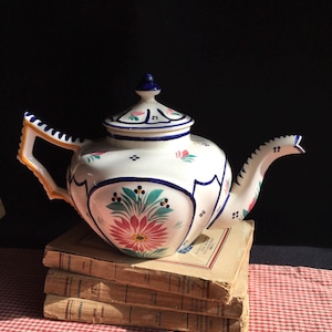 May include: A white ceramic teapot with a floral design in pink, green, and blue. The teapot has a curved spout and handle, and sits atop a stack of old books on a red and white checkered tablecloth.
