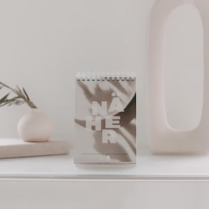 Könnte beinhalten: Ein weißer Schreibtisch mit einer weißen Vase mit getrockneten Blumen, einer kleinen weißen Vase mit einer grünen Pflanze und einem Kalender mit dem aufgedruckten Wort "NÄHER".