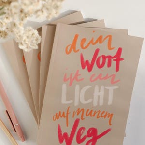 Könnte beinhalten: Ein Stapel beiger Notizbücher mit farbenfrohem handgeschriebenem Text in Orange, Rosa und Weiß. Der Text lautet "dein Wort ist ein Licht auf meinem Weg". Im Hintergrund sind ein paar Stifte und Trockenblumen zu sehen.