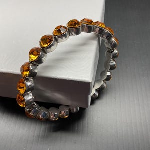 May include: A silver-toned metal bangle bracelet featuring a continuous row of round, faceted amber-colored glass stones set in individual metal bezels. The bracelet has a circular shape and a polished metallic finish, showcasing a series of orange gems.