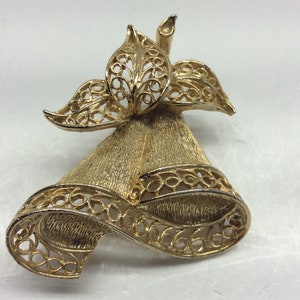 May include: A gold-tone bell-shaped brooch with a filigree design. The bell is decorated with a bow and a floral motif.