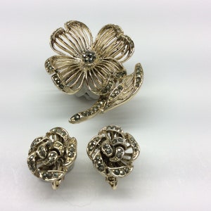 May include: A gold tone flower brooch with a leaf and two matching earrings. The jewelry is set with marcasite stones.