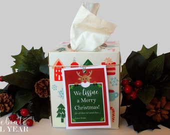 Puede incluir: Una caja de pañuelos blanca con un diseño navideño que presenta casas, copos de nieve y un reno. Una etiqueta roja con una ilustración de reno y el texto "We tissue a Merry Christmas! (for all those red-nosed days.) Love from..." está unida a la caja.