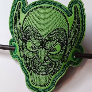 Puede incluir: Parche verde bordado con la cara del Duende Verde con costuras negras. El parche tiene la forma de la cabeza de un duende con orejas grandes.