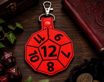 May include: A red, twelve-sided dice keychain with black numbers and a silver clasp. The keychain is designed for role-playing games. Other dice and a leather-bound book are in the background.