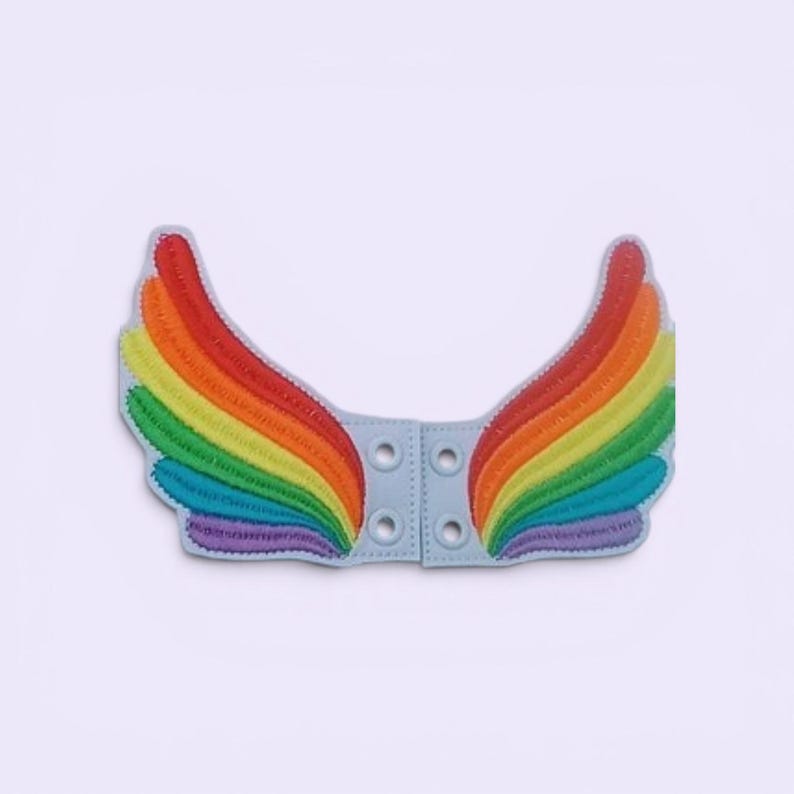 May include: A pair of rainbow-colored embroidered wings with a light blue backing. The wings are stitched with vibrant colors, including red, orange, yellow, green, blue, and purple. The light blue backing has four eyelets for attachment.