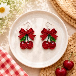 May include: A pair of embroidered cherry earrings featuring red cherries with green leaves and a red bow at the top. Each earring is suspended from a silver-toned metal hook. The design is detailed with textured red and green thread work.