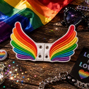 May include: A white leatherette wing-shaped accessory with rainbow embroidery. The image includes a rainbow flag, heart-shaped sunglasses, and a pin with a rainbow heart. A black tag reads "LOVE IS LOVE".