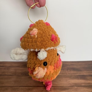 May include: A crocheted mushroom plush keychain featuring a textured brown cap with pink spots, a white stem, and small white wing-like accents. The plush has black bead eyes and is attached to a gold-toned circular metal wire ring for hanging.