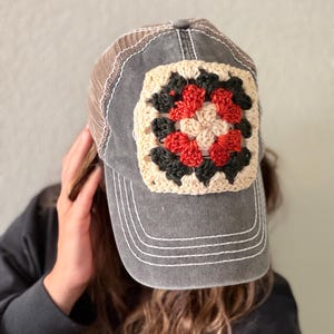 May include: A charcoal gray baseball cap featuring a square crochet granny square patch on the front panel. The patch displays a geometric pattern in cream, black, and rust orange yarn. The cap has a mesh back and white decorative stitching.