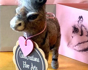 May include: A gray and brown felt donkey with a pink heart and a sign that says "Cun Creations Fiber Arts".