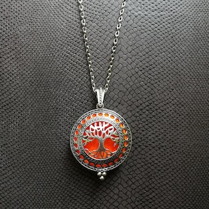 Può includere: Una collana con catena d'argento con un ciondolo rotondo che presenta un design ad albero della vita. L'albero è delineato in argento e riempito con un colore arancione brillante.