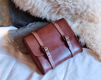 May include: A brown leather satchel with two buckled straps across the front. The bag has a rectangular shape with rounded corners and a flap closure. The leather has a rich, warm tone, and the buckles are a brass color.