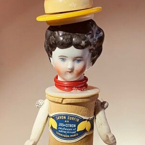 May include: A porcelain doll head with black hair and blue eyes is attached to a wooden spool with a label that reads "Savon Surfin au Jus de Citron onctueux et adoucissant Cosmydor-Paris". The spool is topped with a yellow hat and a yellow and brown hat.