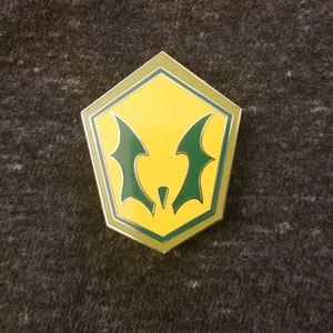 May include: A yellow and green enamel pin with a bat-like design. The pin has a pentagon shape with a silver border. The bat wings are green, and the background is yellow. The pin is on a gray surface.