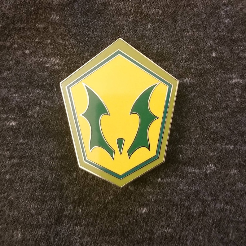 May include: A yellow and green enamel pin with a bat-like design. The pin has a pentagon shape with a silver border. The bat wings are green, and the background is yellow. The pin is on a gray surface.