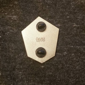 May include: Back of a pentagon-shaped pin with two black pin backs. The pin has a metallic finish and the text "THAT JACK KEEPINGS" is stamped on the surface.