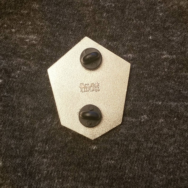 May include: Back of a pentagon-shaped pin with two black pin backs. The pin has a metallic finish and the text "THAT JACK KEEPINGS" is stamped on the surface.