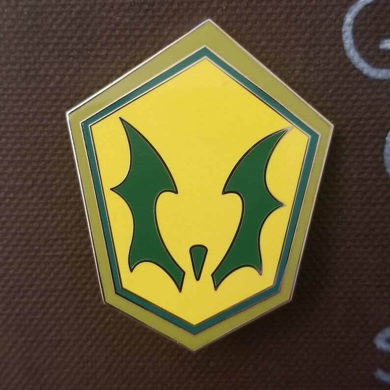 May include: A yellow and green enamel pin with a stylized bat-like design. The pin has a pentagon shape with a green border and a silver outline. The bat wings are green and the background is yellow.