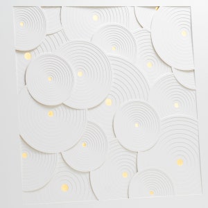May include: White paper art featuring a repeating pattern of concentric circles. The circles are layered and cut out to create a 3D effect. Small gold dots are scattered throughout the design.