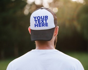 May include: A white and black trucker hat with a black mesh back. The front of the hat has blue text that says "YOUR TEXT HERE".
