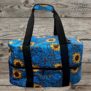 May include: A blue and brown tote bag with a sunflower and floral pattern. The bag has black handles and a zipper closure.