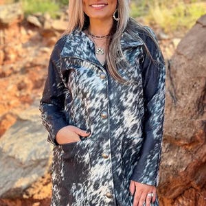 May include: A long black and white cowhide print jacket with a button-up front and two pockets. The jacket has a collar and long sleeves.