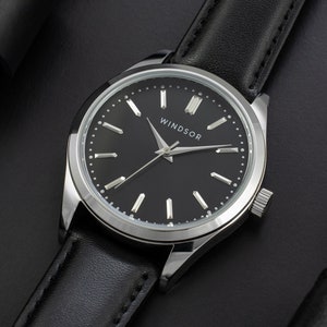 May include: A silver watch with a black leather band and a black face. The watch face has silver hands and markers and the word "WINDSOR" printed on it.