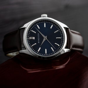 Puede incluir: Un reloj plateado con esfera azul oscuro y correa de cuero marr&oacute;n. La esfera del reloj tiene marcadores plateados y la palabra "WINDSOR". El reloj est&aacute; sobre una superficie marr&oacute;n oscuro.