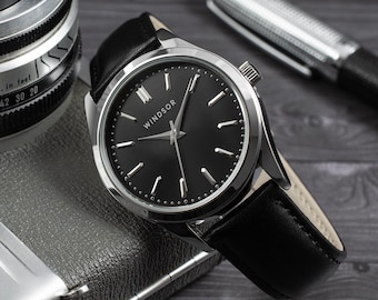 May include: A wristwatch with a round silver-tone case, a black dial featuring the word Windsor, and a black leather strap. The watch has silver-tone hour markers and hands, positioned near a vintage camera lens and a silver pen.