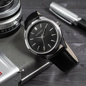 May include: A silver watch with a black face and black leather band. The watch face has silver hands and Roman numeral hour markers. The watch face also has the word "WINDSOR" printed on it. The watch is sitting on top of a silver camera.