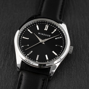 Puede incluir: Un reloj de pulsera plateado con una correa de cuero negro y una esfera negra. La esfera del reloj tiene marcadores de hora en n&uacute;meros romanos plateados y una aguja de segundos plateada. La esfera del reloj tambi&eacute;n tiene la palabra "WINDSOR" impresa en ella.