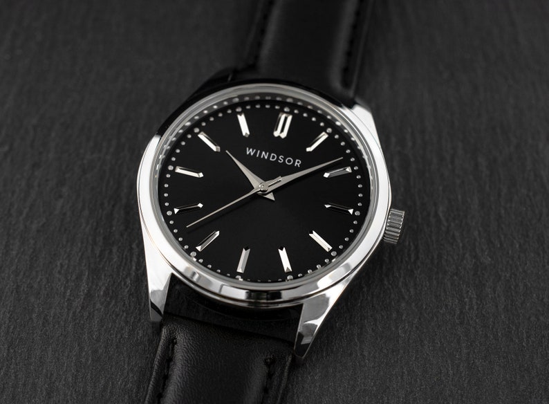 Puede incluir: Un reloj de pulsera plateado con una correa de cuero negro y una esfera negra. La esfera del reloj tiene marcadores de hora en n&uacute;meros romanos plateados y una aguja de segundos plateada. La esfera del reloj tambi&eacute;n tiene la palabra "WINDSOR" impresa en ella.