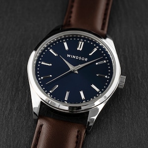 May include: A silver watch with a blue face and brown leather band. The watch face has silver Roman numeral hour markers and the word "Windsor" printed on it.