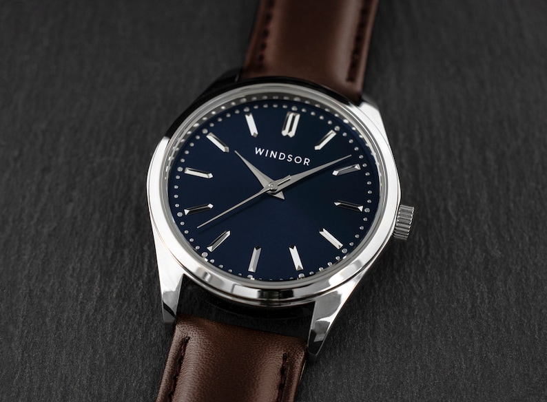 May include: A silver watch with a blue face and brown leather band. The watch face has silver Roman numeral hour markers and the word "Windsor" printed on it.
