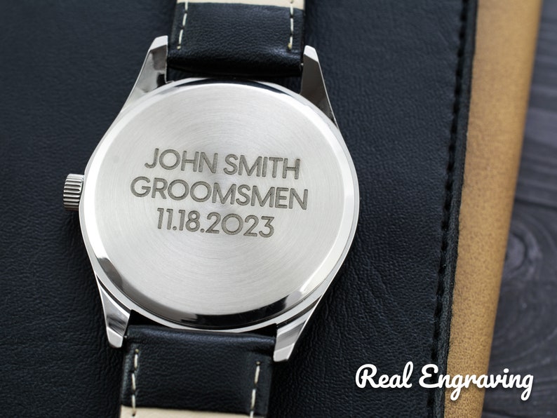 May include: A silver watch with a black leather band. The back of the watch is engraved with the text "JOHN SMITH GROOMSMEN 11.18.2023".