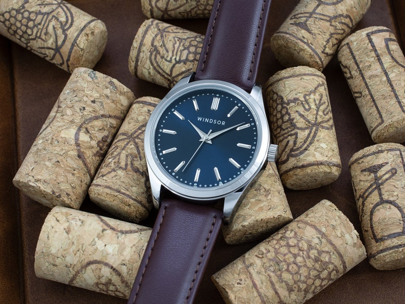 May include: A silver watch with a dark blue face and white hands. The watch has a brown leather band and the word "WINDSOR" printed on the face. The watch is surrounded by cork bottle stoppers.