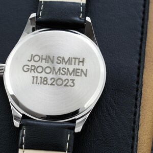 May include: A silver watch with a black leather band. The back of the watch is engraved with the text "JOHN SMITH GROOMSMEN 11.18.2023".