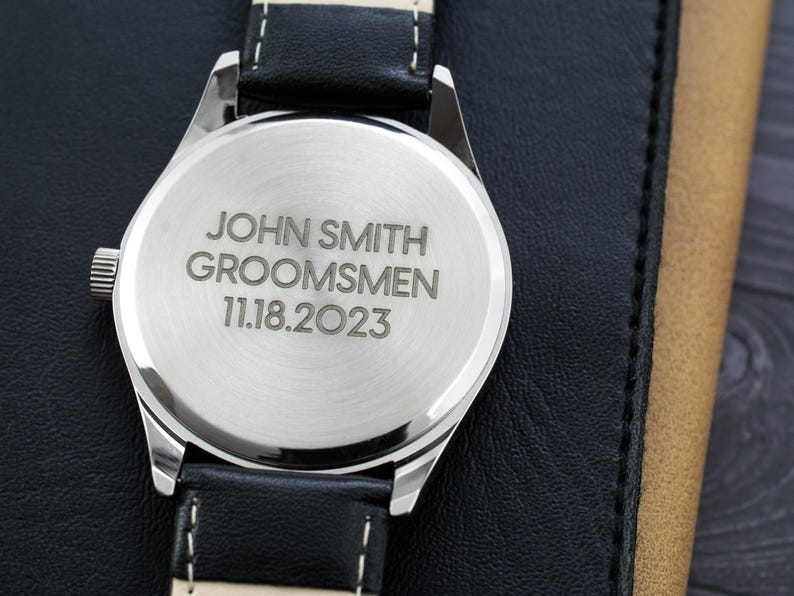 May include: A silver watch with a black leather band. The back of the watch is engraved with the text "JOHN SMITH GROOMSMEN 11.18.2023".