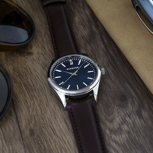 May include: A silver watch with a brown leather band and a blue face. The watch face has silver hands and Roman numeral hour markers. The brand name "WINDSOR" is printed on the watch face.