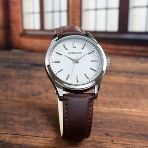 May include: A silver watch with a white face and brown leather band. The watch face has black Roman numeral hour markers and a silver second hand. The watch face also has the word "WINDSOR" printed on it.