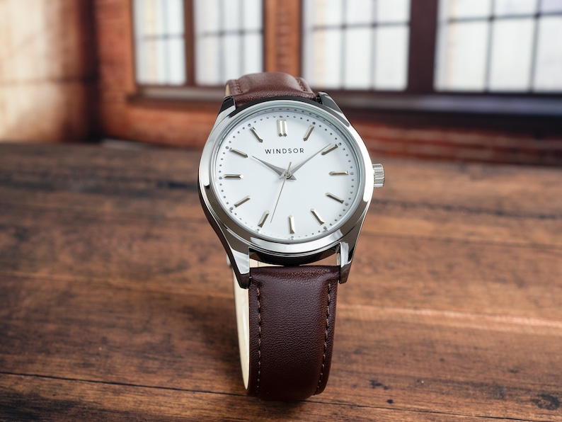 May include: A silver watch with a white face and brown leather band. The watch face has black Roman numeral hour markers and a silver second hand. The watch face also has the word "WINDSOR" printed on it.