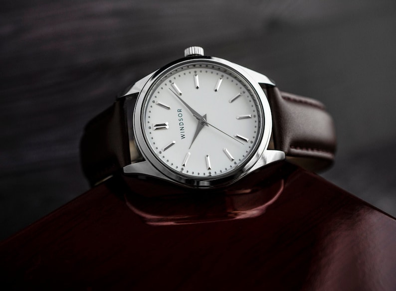 Puede incluir: Un reloj de pulsera plateado con una esfera blanca y marcadores de hora en n&uacute;meros romanos negros. El reloj tiene una correa de cuero marr&oacute;n y la palabra "WINDSOR" est&aacute; impresa en la esfera.