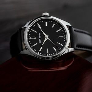May include: A silver wristwatch with a black leather band and a black face with white Roman numeral hour markers and silver hands. The watch face also has the word "WINDSOR" printed on it.
