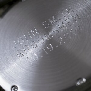 Puede incluir: Primer plano de la parte trasera de un reloj plateado con texto grabado. El texto dice "JOHN SMITH GROOMSMEN 10.19.2019". El reloj tiene un acabado circular de metal cepillado, probablemente un regalo personalizado.