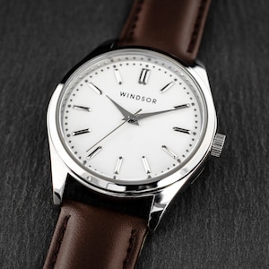 May include: A silver watch with a white face and brown leather band. The watch face has Roman numeral hour markers and the word "WINDSOR" printed on it.