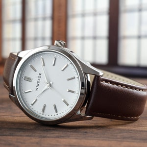 May include: A silver watch with a white face and brown leather band. The watch face has black Roman numeral hour markers and the word "WINDSOR" printed on it.