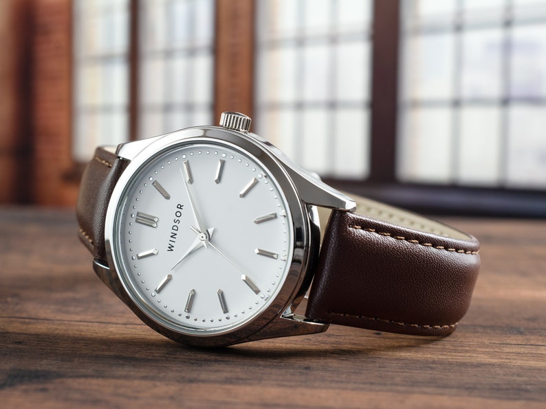 May include: A silver watch with a white face and brown leather band. The watch face has black Roman numeral hour markers and the word "WINDSOR" printed on it.