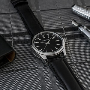 Puede incluir: Un reloj plateado con una correa de cuero negro y una esfera negra. La esfera del reloj tiene agujas plateadas y marcadores de hora en n&uacute;meros romanos. La esfera del reloj tambi&eacute;n tiene la palabra "WINDSOR" impresa.