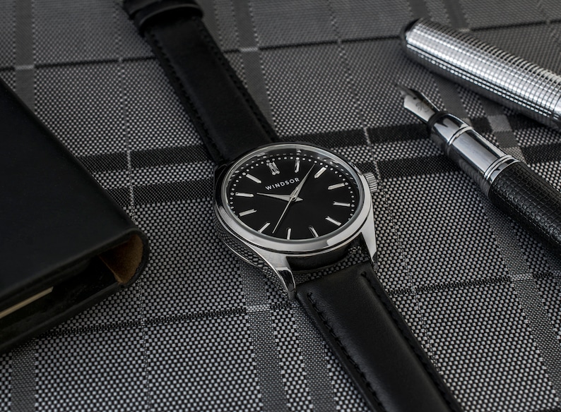Puede incluir: Un reloj plateado con una correa de cuero negro y una esfera negra. La esfera del reloj tiene agujas plateadas y marcadores de hora en n&uacute;meros romanos. La esfera del reloj tambi&eacute;n tiene la palabra "WINDSOR" impresa.